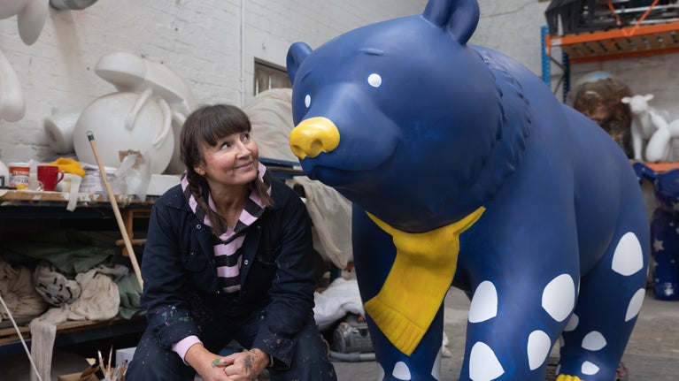 The artist is smiling and looking up at her bear to her left. The big blue bear sculpture is smiling too.
